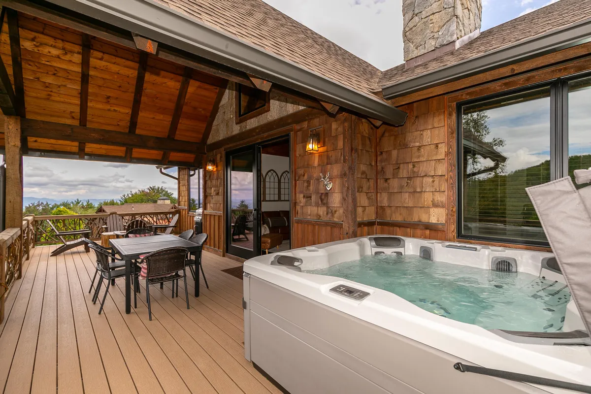 A tranquil exterior scene with a bubbling hot tub, a dining table set for outdoor meals, wooden beams framing the space, and a breathtaking scenic mountain view in the background. Evening ambiance is enhanced by soft lighting, creating a luxurious feel.