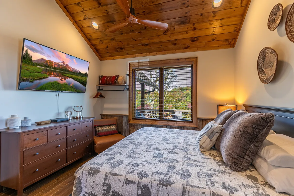 This image features a king-sized bed with a patterned quilt, a large Smart TV displaying a scenic view, a wooden dresser, and a window with blinds that lets in natural light. The setup is cozy and inviting, suggesting a restful place to unwind while enjoying amenities like television and beautiful surroundings.