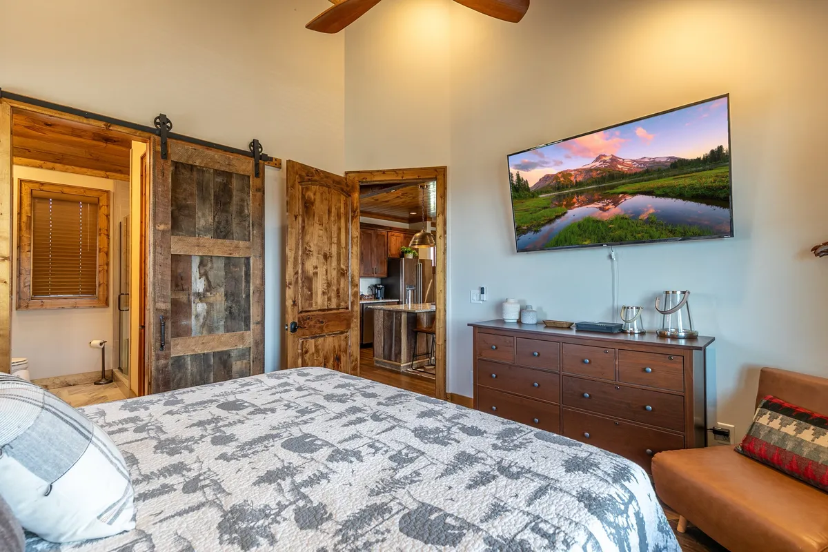This room displays a king-size bed with a patterned quilt, wooden sliding barn doors, a large wall-mounted television, and a wooden dresser. The overall impression is warm and inviting, with a blend of rustic charm and modern convenience.