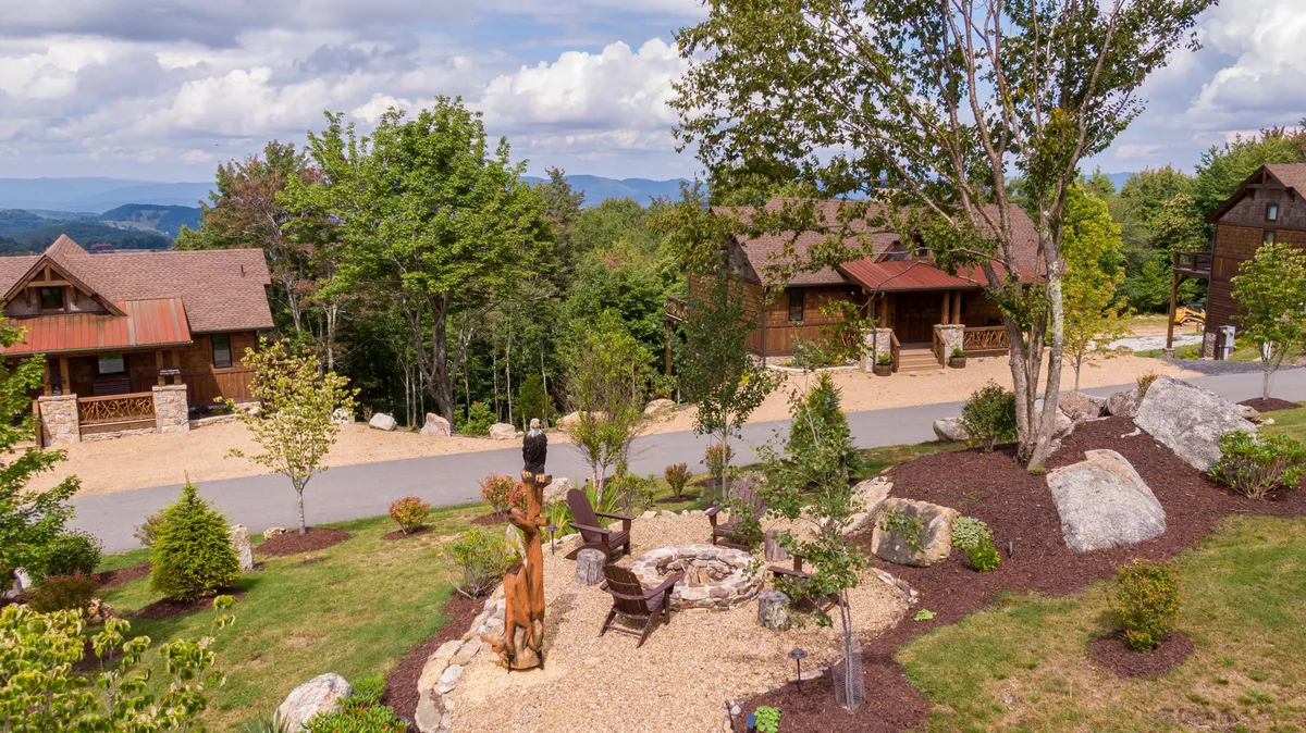 This image captures a group-friendly exterior space featuring a stone firepit surrounded by chairs, set amid tall trees and classic cabins. The setting is ideal for socializing, sharing stories around the fire, or relaxing after a day of activities. The presence of cabins in the background adds to the communal, retreat-like feeling.