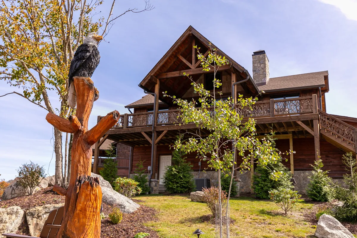 A wide exterior view of the home’s log cabin architecture, accented by a striking wooden eagle sculpture, lush greenery, and rugged stone details. The setting is rich with natural elements, suggesting proximity to woodland trails and opportunities for nature walks and wildlife watching.