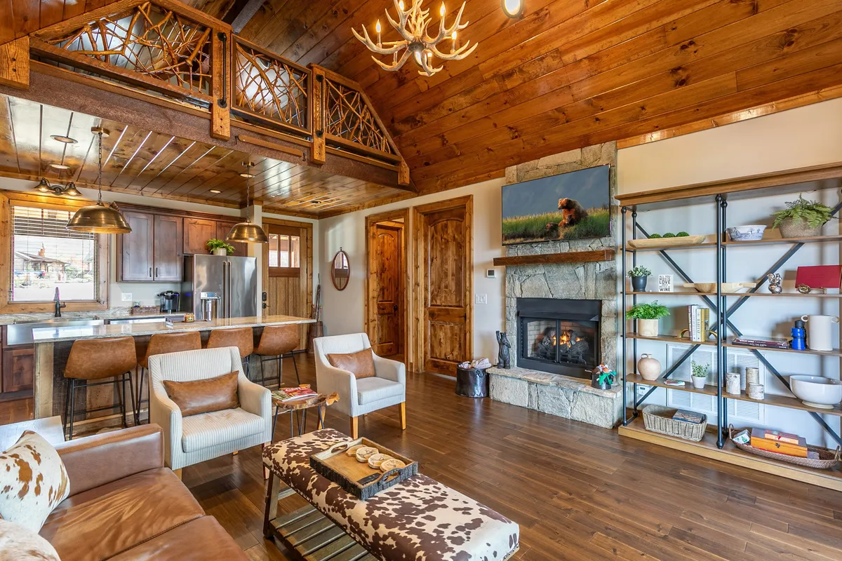A wide shot of the open-concept living area showcasing the fireplace, wooden beams overhead, an open kitchen with modern appliances, and a cozy sofa with a cowhide pattern. Decorative shelving and thoughtful design touches emphasize the space’s blend of comfort and style, making it perfect for group activities and shared meals.