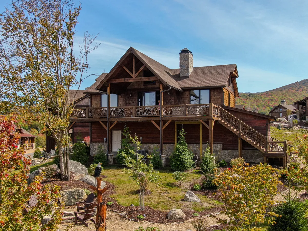 The exterior of a large wooden house with a prominent stone chimney, surrounded by trees and lush landscaping. A balcony offers an elevated spot for enjoying panoramic views and stargazing. The scene conveys a sense of seclusion, luxury, and natural beauty—perfect for a romantic retreat.