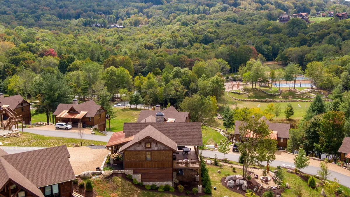 A sweeping view across the resort community, featuring several rustic wooden cabins set among open fields, meandering paths, lush greenery, and water features. The image highlights families and friends gathering for outdoor games, picnics, and walks, with ample space for recreational activities and scenic enjoyment.