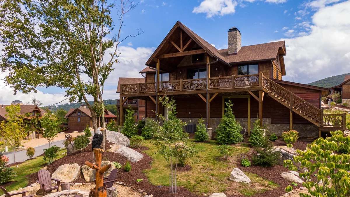 The image presents a spacious wooden deck with a stone chimney, landscaped gardens, and rock formations, offering an inviting space for outdoor dining and gatherings. The deck is oriented toward scenic mountain and garden views, with seating that encourages group interaction.