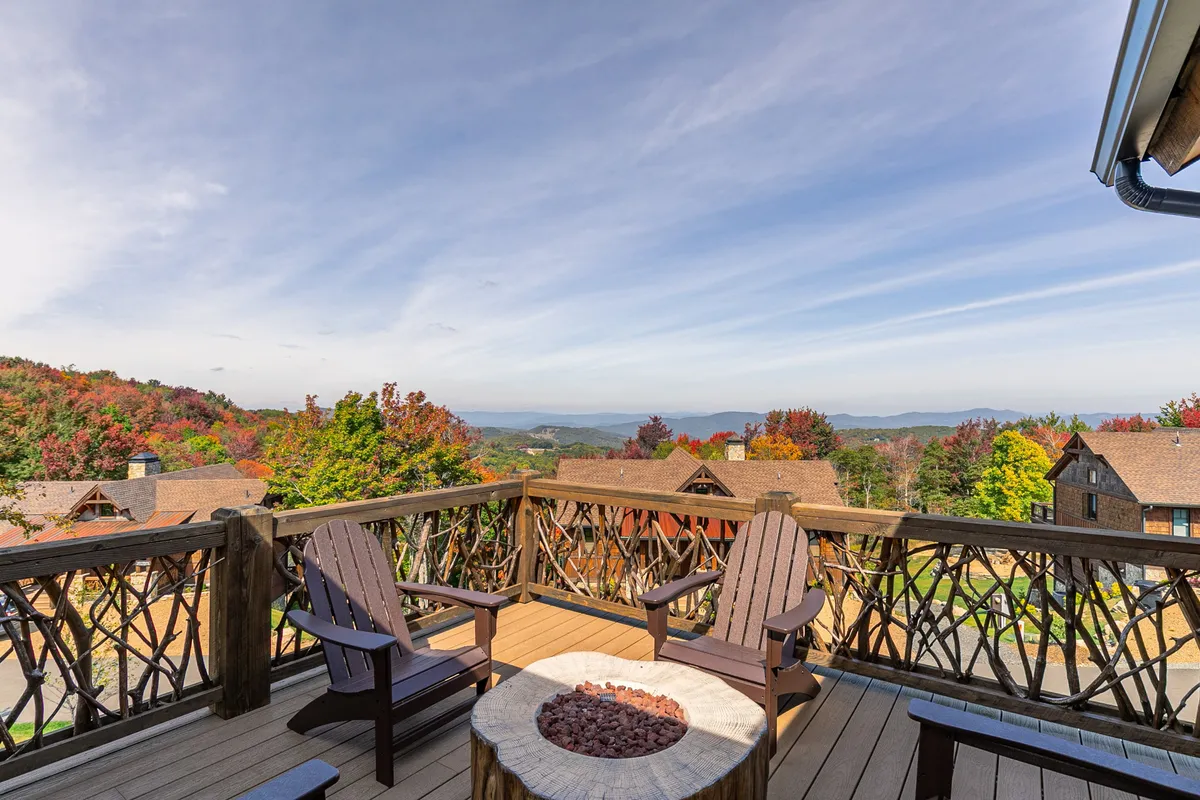 A spacious wooden deck overlooking a breathtaking mountain view at sunset. The deck features a fire pit surrounded by comfortable seating, with lush trees framing the scene. The tranquil ambiance invites relaxation, stargazing, and intimate moments outdoors.