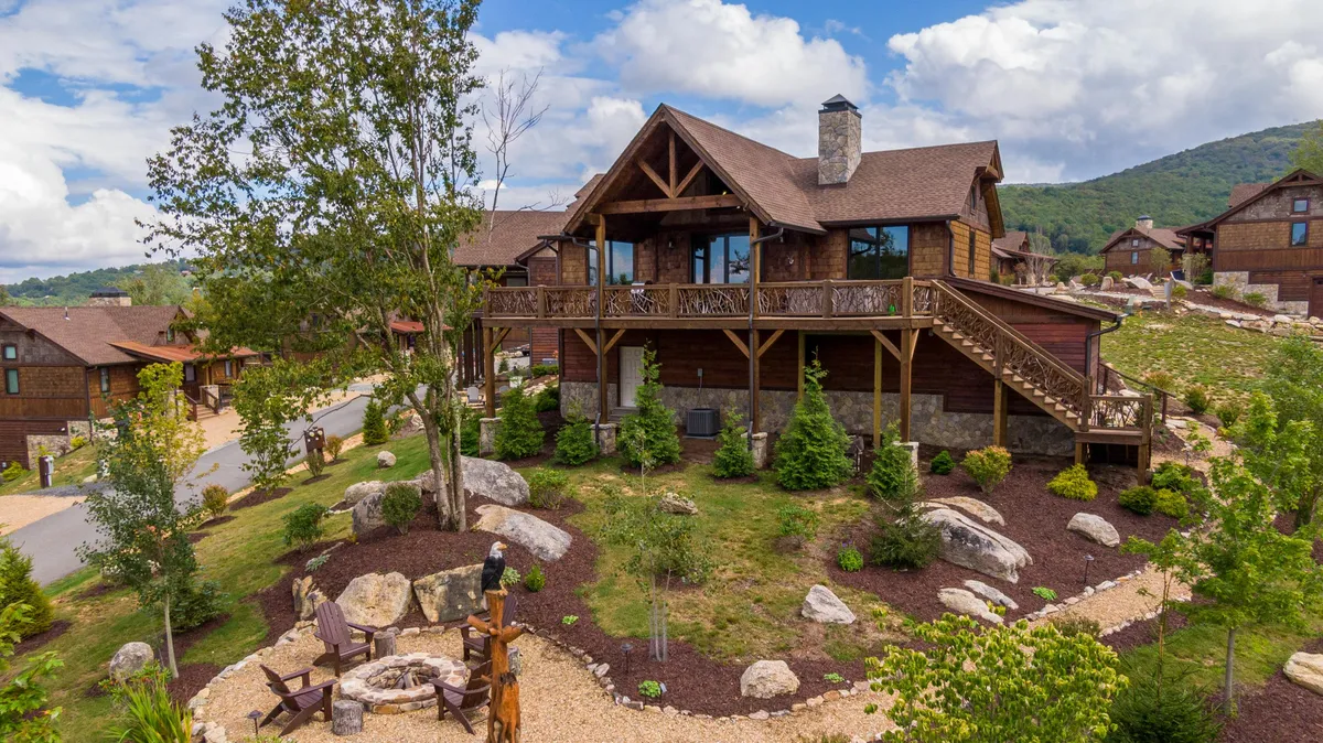 An inviting exterior view of a wooden cabin surrounded by lush landscaping and a cozy fire pit area. The scene shows residents gathering around the fire pit, relaxing on the deck, and enjoying the tranquility of nature, with trails and open space nearby for exploration.