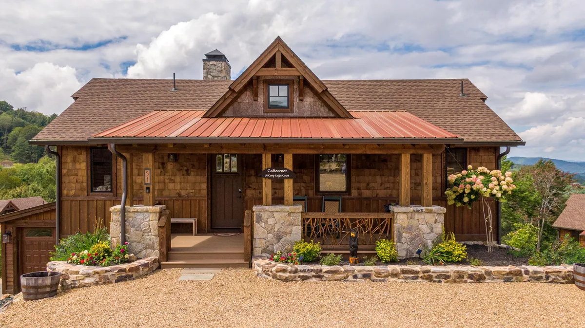 A welcoming exterior shot of the home, complete with a front porch supported by wooden beams, a stone façade, red metal roof, and vibrant flower planters. The porch beckons relaxation and socializing, surrounded by mountain scenery and natural beauty.