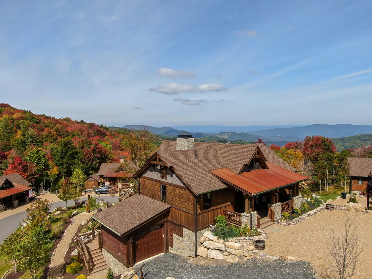 The image showcases a charming single-family mountain home nestled amidst vibrant autumn foliage, with a sweeping backdrop of Blue Ridge Mountains. The spacious back deck faces panoramic views on both sides, while a stone pathway leads guests through the colorful landscape, inviting outdoor relaxation and adventure. The serene morning light hints at the promise of both breathtaking sunrises and sunsets.
